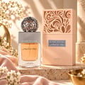 Lattafa Beyond Precious by Lattafa 100ml EDP Women — Botol minyak wangi kaca jernih berisi cecair oren dan penutup perak berhias, diletakkan bersebelahan kotak berwarna pic senada dengan ukiran menarik. Kain merah jambu lembut dan bunga putih mengelilingi paparan.