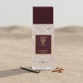 Bujairami Billion Dollar by Bujairami 100ml Extrait — Clear glass perfume bottle with a maroon cap, set on sand with a vanilla bean and sandalwood pieces.
