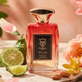 Bujairami Blush Eau de Parfum — A red-toned glass perfume bottle with a gold cap, surrounded by bergamot fruits, sliced lime, almonds, and pink flowers.