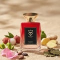 Bujairami Boujee by Bujairami 100ml EDP — Red faceted glass bottle with gold cap, surrounded by lychees, ginger slices, lime zest, rose petals, and cocoa beans on a sandy background.