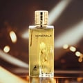 Maison Alhambra Minerale Gold by Maison Alhambra 100ml EDP — A tall, rectangular gold-colored perfume bottle with a fluted gold cap, standing on a reflective surface against a blurred, warm-toned background with bokeh lights.