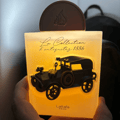 Lattafa Pride La Collection 1886 (Car) — A glowing, translucent bottle with a vintage car silhouette and Arabic script, held in a hand against a dark background.