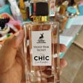 Maison Alhambra Chic Velvet Pink Secret for Women 100ml — A hand holds a clear glass perfume bottle with a white label featuring a deer logo, the text "MAISON ALHAMBRA VELVET PINK SECRET CHIC Eau de parfum" and a heart design. | Maison Alhambra Chic Velvet Pink Secret for Women 100ml — Tangan memegang botol minyak