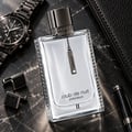 Armaf Club de Nuit Precieux II — Rectangular clear glass perfume bottle with metallic patterned trim, silver tag, on a dark marble surface with a watch, cufflinks, and pen.