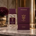 Bujairami Dead Serious by Bujairami 100ml Extrait Unisex — Clear faceted glass perfume bottle with a burgundy cap and label, next to its matching burgundy presentation case on a marble surface, with a dark red flower in the background.