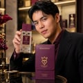 Bujairami Dead Serious by Bujairami 100ml Extrait Unisex — Man holding a clear glass perfume bottle with a burgundy cap and gold label, next to a burgundy leather case with gold branding.