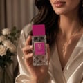 Bujairami First Class Amber Woody 100ml Extrait — Woman holding a clear, faceted glass perfume bottle with a pink cap and a gold and pink label, with white roses in the background.