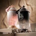 Zimaya Ghali Elegant Eau De Parfum 100ml by Zimaya — Two ornate perfume bottles, one pink and one black, with tassels and gold accents, displayed on a reflective surface against a textured beige background. | Zimaya Ghali Elegant Eau De Parfum 100ml by Zimaya — Dua botol minyak wangi berhias, satu merah jambu dan satu