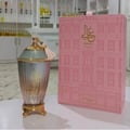 Zimaya Ghali Elegant Eau De Parfum 100ml by Zimaya — A tall, fluted glass perfume bottle with a gold base and a light blue textured cap adorned with a pink tassel, stands next to its matching pink box decorated with a building facade pattern. | Zimaya Ghali Elegant Eau De Parfum 100ml by Zimaya — Botol kaca berlekuk