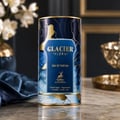 Maison Alhambra Glacier Flora 100ml — A tall, cylindrical perfume bottle with a dark blue and gold floral design, set against a dark background with blurred white flowers and gold accents. | Maison Alhambra Glacier Flora 100ml — Botol minyak wangi silinder tinggi dengan reka bentuk bunga biru gelap dan emas