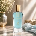 Maison Alhambra Glacier Flora 100ml by Maison Alhambra — A clear glass perfume bottle with light blue liquid and a gold cap, featuring gold text and a deer logo, sits on a marble surface next to a vase of white flowers and draped blue fabric. | Maison Alhambra Glacier Flora 100ml by Maison Alhambra — Botol kaca jernih