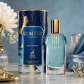 Maison Alhambra Glacier Flora — A blue and gold cylindrical box stands next to a clear glass bottle of light blue perfume, both featuring gold deer motifs and text. A white flower and delicate white blossoms are visible in the background. | Maison Alhambra Glacier Flora — Kotak silinder biru dan emas berdiri di