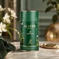 Maison Alhambra Glacier Heaven 100ml — A tall, dark green cylindrical perfume bottle with gold trim and floral and bird illustrations, placed on a marble surface with a green silk cloth and a gold tray. | Maison Alhambra Glacier Heaven 100ml — Botol minyak wangi silinder hijau gelap tinggi dengan trim emas dan