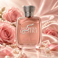 Kenz de Rasasi Hawas Desire First Kiss — Botol minyak wangi rose gold dengan lambang ular perak dan penutup perak berhias, dikelilingi bunga ros merah jambu dan mutiara di atas permukaan marmar.