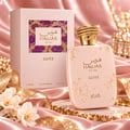 RASASI Hawas Glitz by Rasasi 100ml EDP — A light pink perfume bottle with gold speckles and a textured gold cap, placed next to its matching pink box with a purple and gold patterned insert, on a pink satin background with white flowers and gold ribbon.