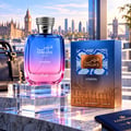 RASASI Hawas London by Rasasi 100ml EDP — Botol minyak wangi gradien biru dan merah jambu dengan penutup perak seperti ular terletak di atas tapak akrilik jernih, bersebelahan kotak birunya yang menampilkan corak ular dan ilustrasi langit bandaraya London. Beg kulit gelap dan pasport biru turut kelihatan.