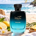 RASASI Hawas Malibu by Rasasi 100ml EDP — A sleek, gradient blue and black perfume bottle with gold Arabic script and "HAWAS For Him MALIBU" text, set on a wooden surface with lemons, a plumeria flower, and pineapple slices, with a blurred beach and ocean background.