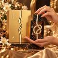 Maison Alhambra IT by Maison Alhambra 100ml EDP Men — Kotak emas dan botol minyak wangi rekaan simpulan emas, dipegang tangan bertindik cincin dan gelang, dikelilingi bunga dan fabrik mewah.