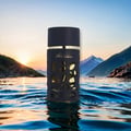Lattafa Khalta Blend Of Lattafa Eau De Parfum 100ml — Botol minyak wangi gelap dengan rekaan berlubang dan kemasan emas, terapung di atas air dengan latar belakang gunung dan matahari terbenam.