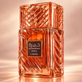 Lattafa Khamrah Mira — Botol minyak wangi kaca ton ambar dengan corak geometri rumit, berlatar belakang fabrik lembut berlipat dengan lilin menyala, kayu manis, ketulan coklat, dan beri merah.