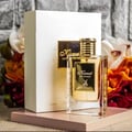Maison Alhambra Kismet for Women 100ml by Maison Alhambra — A clear glass perfume bottle with a gold cap and gold label featuring a deer silhouette, placed next to its white and gold box, with blurred floral arrangements in the background. | Maison Alhambra Kismet for Women 100ml by Maison Alhambra — Botol minyak
