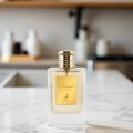 Maison Alhambra Kismet for Women 100ml by Maison Alhambra — A clear glass perfume bottle with a gold fluted cap and a gold label featuring a deer silhouette, placed on a marble countertop with a blurred kitchen background. | Maison Alhambra Kismet for Women 100ml by Maison Alhambra — Botol minyak wangi kaca jernih