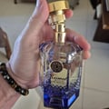 Maison Alhambra Kismet Lunar Magic 100ml by Maison Alhambra — Hand holding a clear faceted glass perfume bottle with blue liquid and gold accents, showing the cap being twisted off. | Maison Alhambra Kismet Lunar Magic 100ml by Maison Alhambra — Tangan memegang botol minyak wangi kaca berfaset jernih dengan cecair