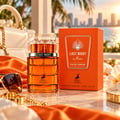 Maison Alhambra Last Night in Miami — Amber glass perfume bottle and orange box displayed with a white handbag, sunglasses, and gold jewelry on a marble surface, with a city skyline and palm trees in the background.