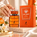 Maison Alhambra Last Night in Miami — A hand with a gold bracelet holds a clear glass perfume bottle with amber liquid. The bottle has a silver band and the box is orange.