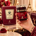 Maison Alhambra Last Night in Paris — A hand holds a red glass perfume bottle with a gold cap, next to its matching red box, on a marble surface with pearls and a rose petal.
