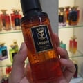 Bujairami Legacy by Bujairami 100ml Extrait — Hand holding an amber glass perfume bottle with a black cap and a black label featuring a gold crown logo and the text 'Legacy'. Several other perfume bottles are visible on shelves in the background.
