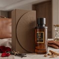 Bujairami Mad Love 100ml Extrait Unisex — A brown glass perfume bottle with a black cap and gold label, next to its matching brown textured box. Rose petals, vanilla beans, and wood pieces are arranged around the bottle and box on a marble surface.