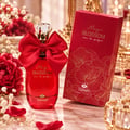 TADangel Magic Blossom — Red glass perfume bottle with a large red satin bow, crystal cap, and gold accents, next to its red floral-patterned box on a marble surface with red roses and baby's breath.