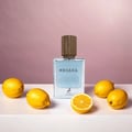 Maison Alhambra Megara 50ml by Maison Alhambra — A clear glass perfume bottle with a wooden cap and blue liquid, surrounded by whole and sliced lemons on a white surface against a pink gradient background. | Maison Alhambra Megara 50ml by Maison Alhambra — Botol minyak wangi kaca jernih bertudung kayu dan cecair biru