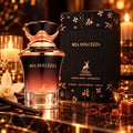 Maison Alhambra Mia Dolcezza by Maison Alhambra 100ml EDP Women — A dark gradient perfume bottle with a rose gold cap, next to its black patterned box, with vanilla pods, a white flower, lit candles, and chocolate pieces in the background.