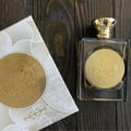 Ard Al Zaafaran Mithqal 100ml by Ard Al Zaafaran — A clear glass perfume bottle with a gold embossed circular label and a gold cap sits next to its matching white and gold patterned box on a dark wooden surface. | Ard Al Zaafaran Mithqal 100ml oleh Ard Al Zaafaran — Botol minyak wangi kaca jernih dengan label bulat