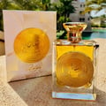 Ard Al Zaafaran Mithqal 100ml — A square glass perfume bottle with a gold emblem and golden liquid sits next to its matching white and gold patterned box, set against a blurred background of water and palm trees. | Ard Al Zaafaran Mithqal 100ml — Sebotol minyak wangi kaca berbentuk segi empat dengan lambang emas dan