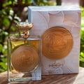 Ard Al Zaafaran Mithqal 100ml — A square glass perfume bottle with a gold cap and a circular gold emblem on the front, placed next to its white and gold patterned box, with a blurred green leafy background. | Ard Al Zaafaran Mithqal 100ml — Botol minyak wangi kaca segi empat dengan penutup emas dan lambang emas bulat