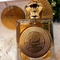 Ard Al Zaafaran Mithqal 100ml — Close-up of a golden perfume bottle with intricate Arabic calligraphy on a circular emblem, set against a soft white fluffy background with a hint of greenery. | Ard Al Zaafaran Mithqal 100ml — Pandangan dekat botol minyak wangi keemasan dengan kaligrafi Arab yang rumit pada lambang