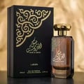 Lattafa Musk Al Aroos by Lattafa 80ml EDP — A black perfume box with ornate gold Arabic calligraphy and scrollwork is placed next to a clear glass perfume bottle with amber liquid and a gold cap. | Lattafa Musk Al Aroos by Lattafa 80ml EDP — Kotak minyak wangi hitam dengan kaligrafi Arab emas yang berhias dan ukiran