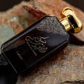 Lattafa Musk Al Aroos by Lattafa 80ml EDP — A dark glass perfume bottle with gold ornate detailing and a gold cap rests on textured rocks. | Lattafa Musk Al Aroos by Lattafa 80ml EDP — Botol minyak wangi kaca gelap dengan perincian berukir emas dan penutup emas diletakkan di atas batu bertekstur.