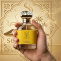 Bujairami Next Level by Bujairami 100ml Extrait — Hand holding a clear glass perfume bottle with a yellow textured band and a crystal-like cap, against a golden desert-themed background with palm trees and ornate patterns.