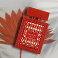 Rave Now Rouge Perfume EDP 100ml by RAVE — A red perfume bottle with a geometric cut-out design and a red cap sits next to a dried palm leaf on a light blue background with shadows. | Rave Now Rouge Perfume EDP 100ml by RAVE — Botol minyak wangi merah dengan reka bentuk potongan geometri dan penutup merah terletak di