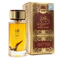 Ard Al Zaafaran Oud Al Turas — A clear glass perfume bottle with golden cap and a dark brown dried plant inside, next to its ornate gold and maroon box. | Ard Al Zaafaran Oud Al Turas — Botol minyak wangi kaca jernih bertutup emas dan tumbuhan kering coklat gelap di dalamnya, bersebelahan dengan kotak emas dan merah