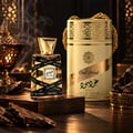 Lattafa Oud Mood 30ml by Lattafa EDP Unisex — A clear glass perfume bottle with a gold cap, wrapped in black ribbon with 'Oud Mood' text, sits on a wooden block. A matching cylindrical gold and cream presentation box is behind it, with smoke rising from the left.