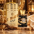 Lattafa Oud Mood 30ml by Lattafa EDP Unisex — A tall, cream-colored cylindrical box with gold ornate patterns and a gold chain handle, next to a clear glass perfume bottle with a gold cap, wrapped in black ribbon printed with 'Oud Mood'.