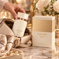 Khadlaj Panache Angel Dust — A hand holding a cream-colored perfume bottle with gold accents, placed inside a cream handbag. A matching perfume box sits beside it, with floral embossed details.
