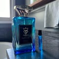 Bujairami Perfect Life by Bujairami Eau de Parfum 100ml — Blue glass perfume bottle with a silver cap, next to a small sample vial on a dark countertop with a blurred background of a bathroom.