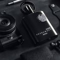 Afnan Supremacy Noir by Afnan 100ml EDP Men — Botol wangian hitam legam dengan logo perak, diletakkan di atas permukaan gelap bersama kamera vintaj, dompet kulit, dan fon telinga