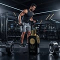 Ard Al Zaafaran Dirham Gold By Ard Al Zaafaran 100ml For Him — Botol wangian hitam dengan penutup emas terletak di atas permukaan gelap berair, dengan seorang lelaki berotot mengangkat dumbbell di gimnasium yang diterangi lampu neon di latar belakang